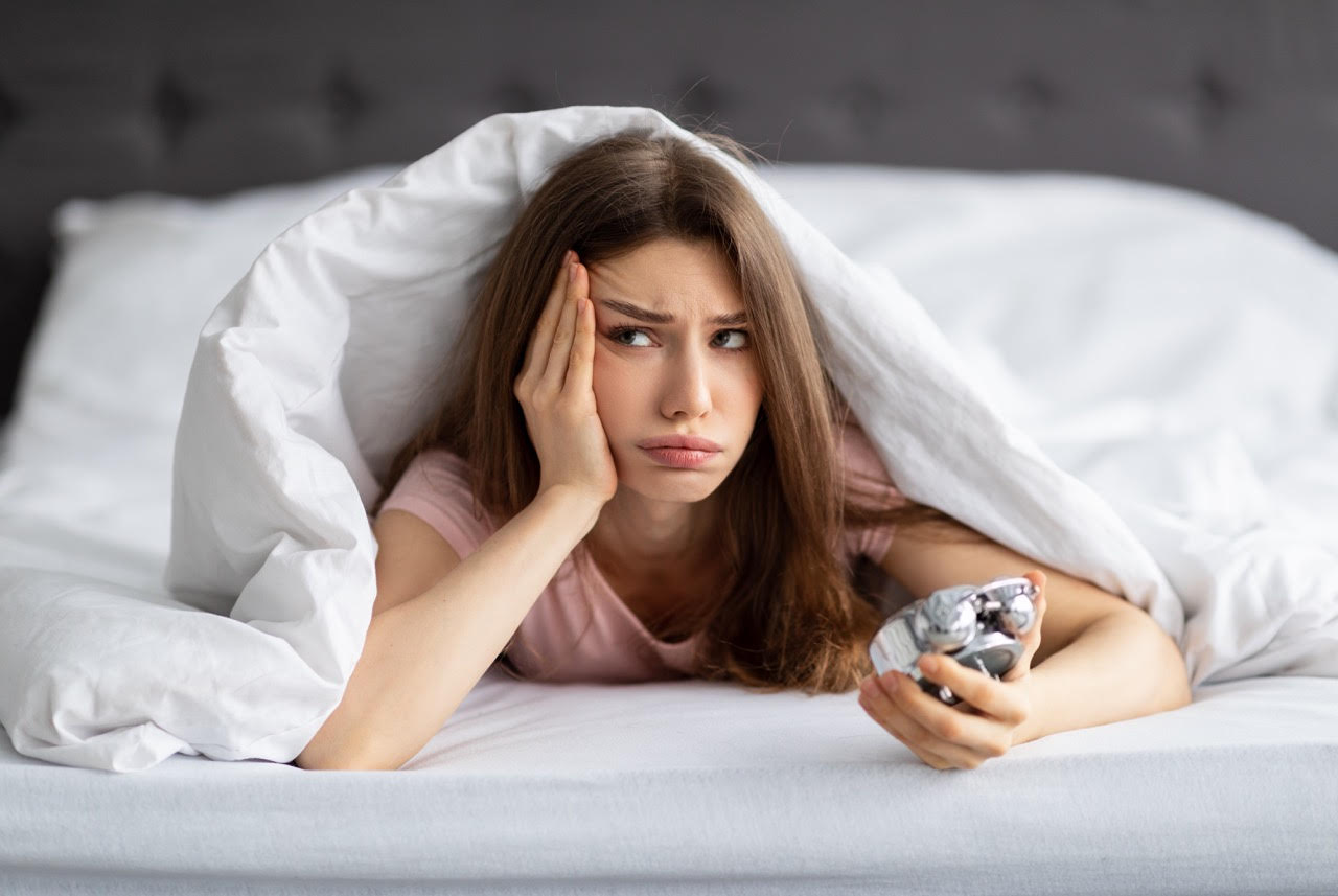 Unhappy young Caucasian woman lying in bed with alarm clock, not willing to get up in morning. Upset millennial lady having problem waking up after sleepless night, cuddling under warm blanket